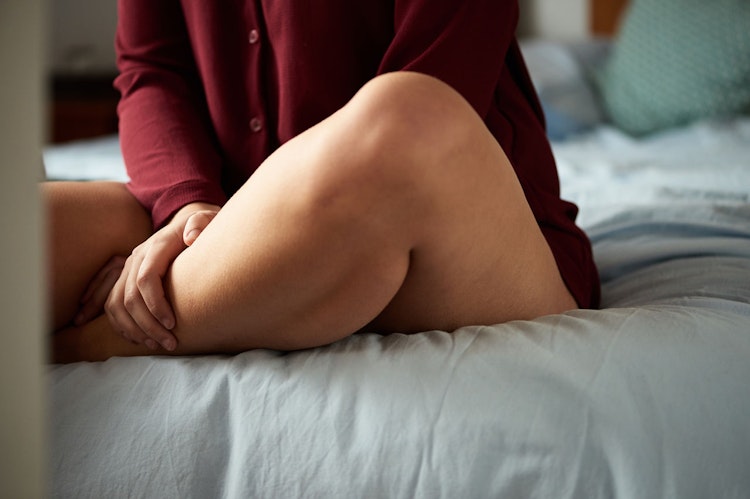 Woman sat on the end of her bed, sitting cross-legged holding her bare legs.