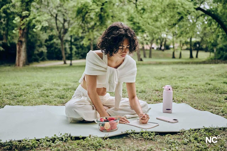 Photograph of a woman sat in a park wearing loose white clothes, journalling with a bowl of fruit, water bottle and her phone beside her.