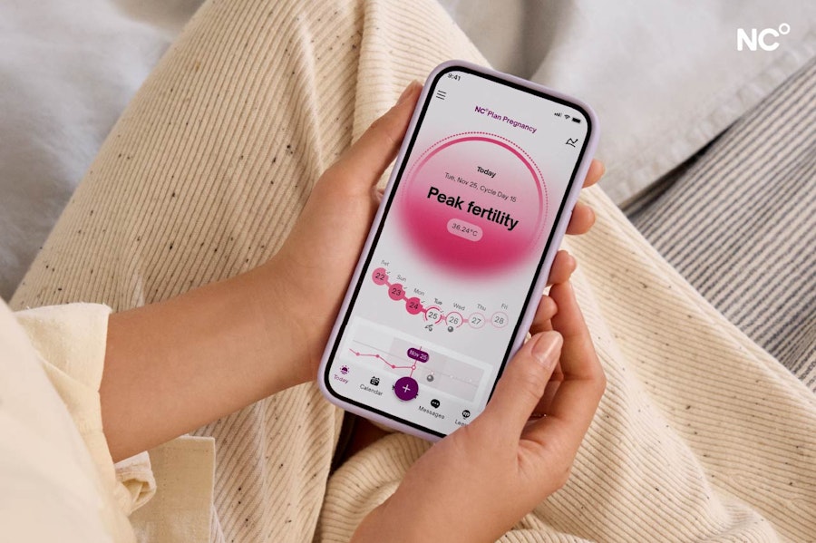 Photograph of hands holding a phone showing the Natural Cycles plan pregnancy app screen with the text "peak fertility"