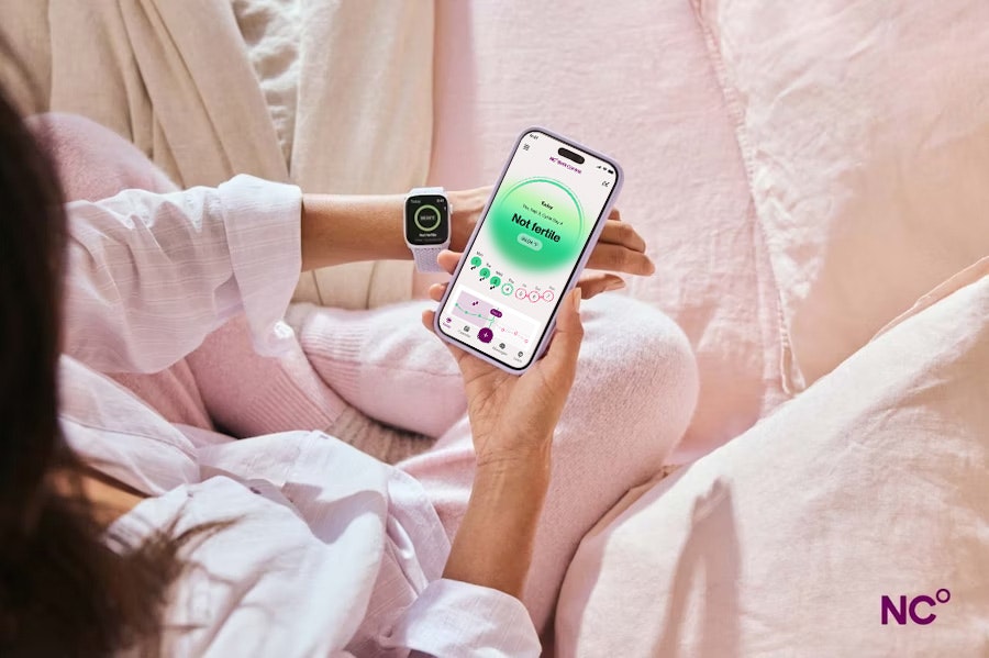 View looking over a woman's shoulder as she sits in bed holding her phone showing the Natural Cycles app with the text 'Not fertile', she's wearing sleepwear and an Apple Watch on her left wrist.