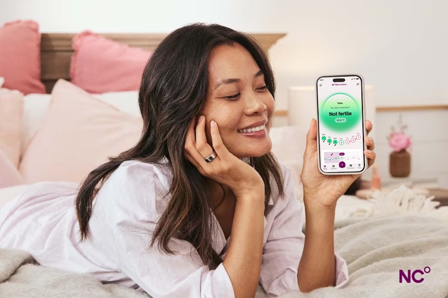 Woman lying on her bed showing the Natural Cycles app on her phone with the text 'Not fertile' on her right ring finger she wears an Oura Ring.