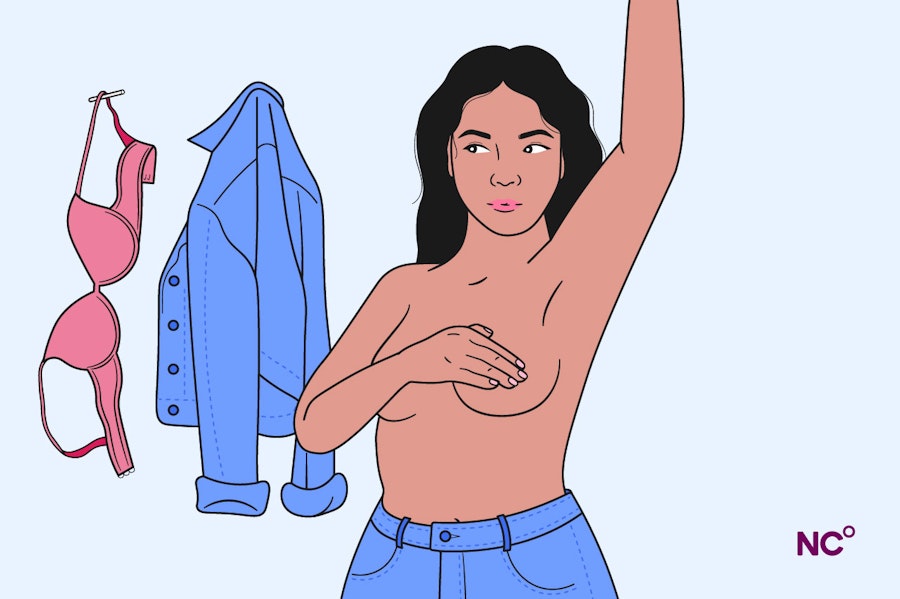 Woman with her bra and shirt hanging up behind her touching her breast with her hand to check for lumps and changes, her other arm is raised in the air