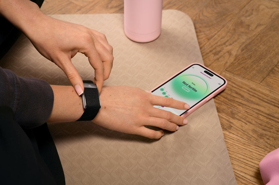 Photograph of a woman's hands on a yoga mat. On her right wrist she wears a WHOOP Band and is holding a phone in a pink case showing the Natural Cycles app with the text Not Fertile. Just at the side of the shot is a pink weight and pink waterbottle.