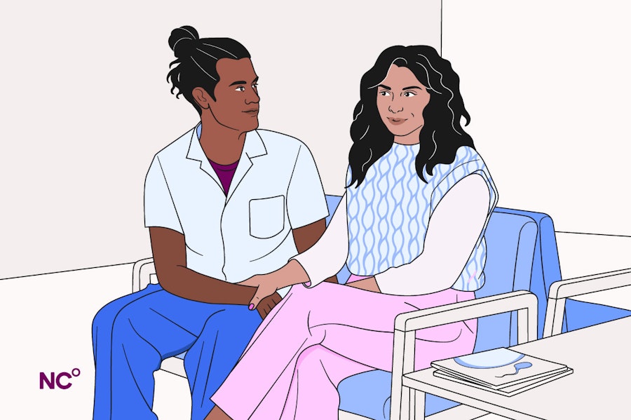 Couple sitting in a medical waiting room, they are looking at each other and holding hands.