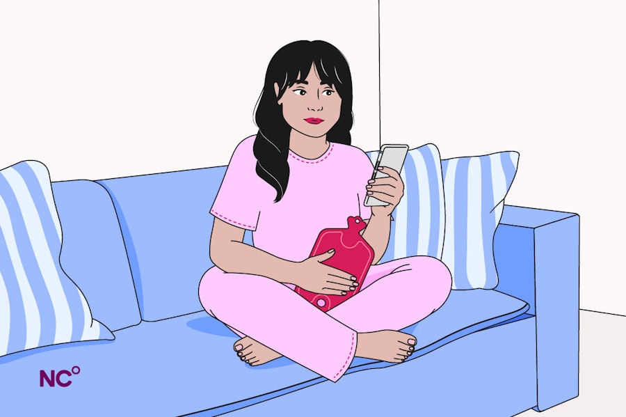 Woman sat on the sofa in her pjs with a hot water bottle holding her phone