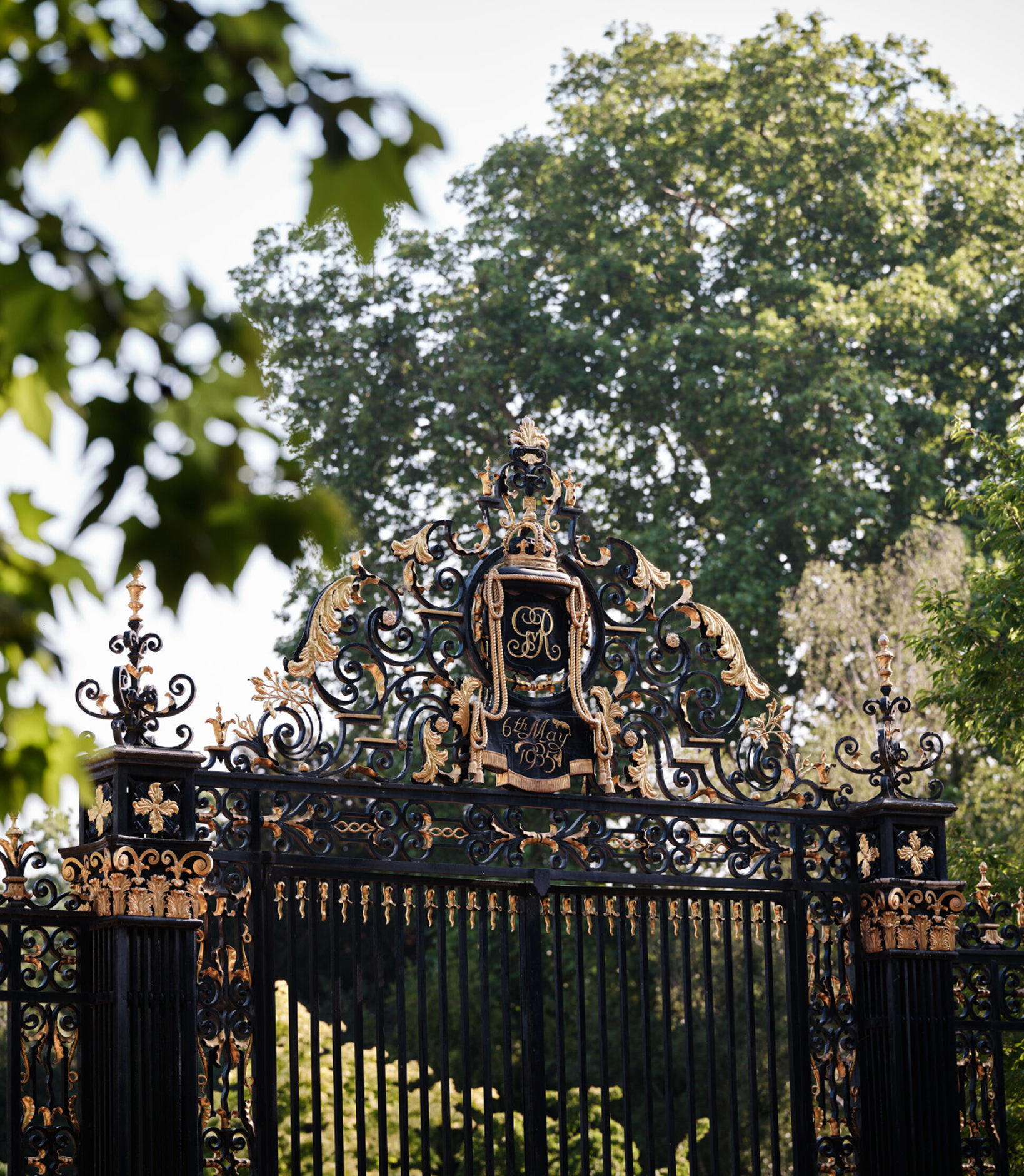 Ornate black and gilded park gates with a royal cypher, framed by tall trees in the Regent's Park