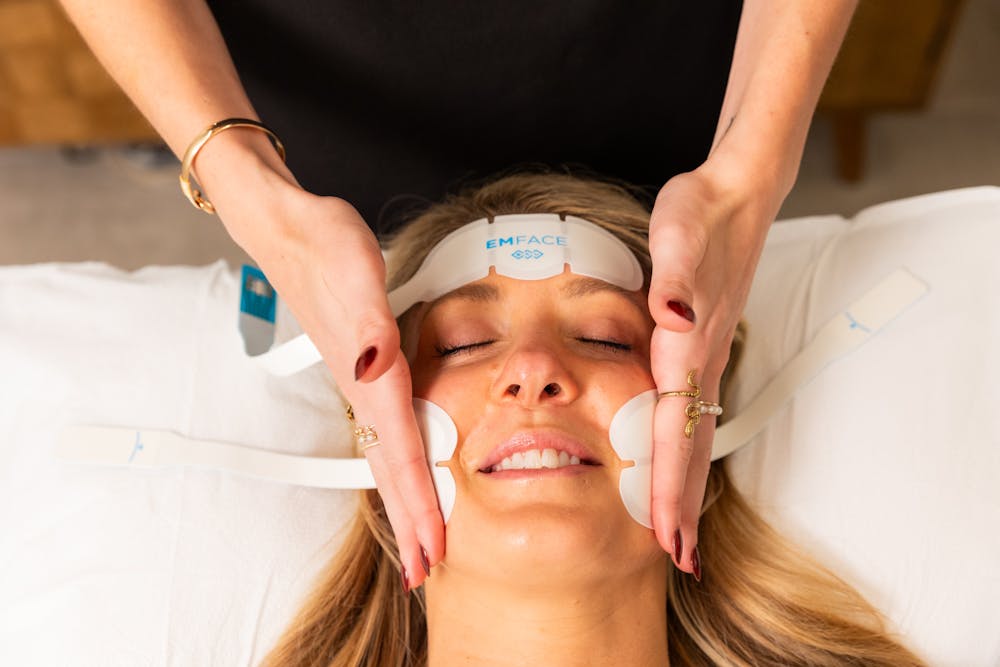 A young blonde female guest smiles, relaxing on a treatment table, as a Lume provider places the Emface cheek applicators for a non-surgical facelift in Chicago.