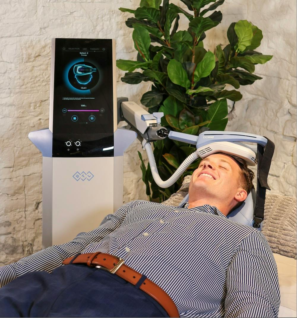 patient lying on back and smiling during a PTSD treatment in Chicago. There is an ExoMind computer screen placed behind him, and an applicator rests comfortably on his head to reactivate his neural pathways.