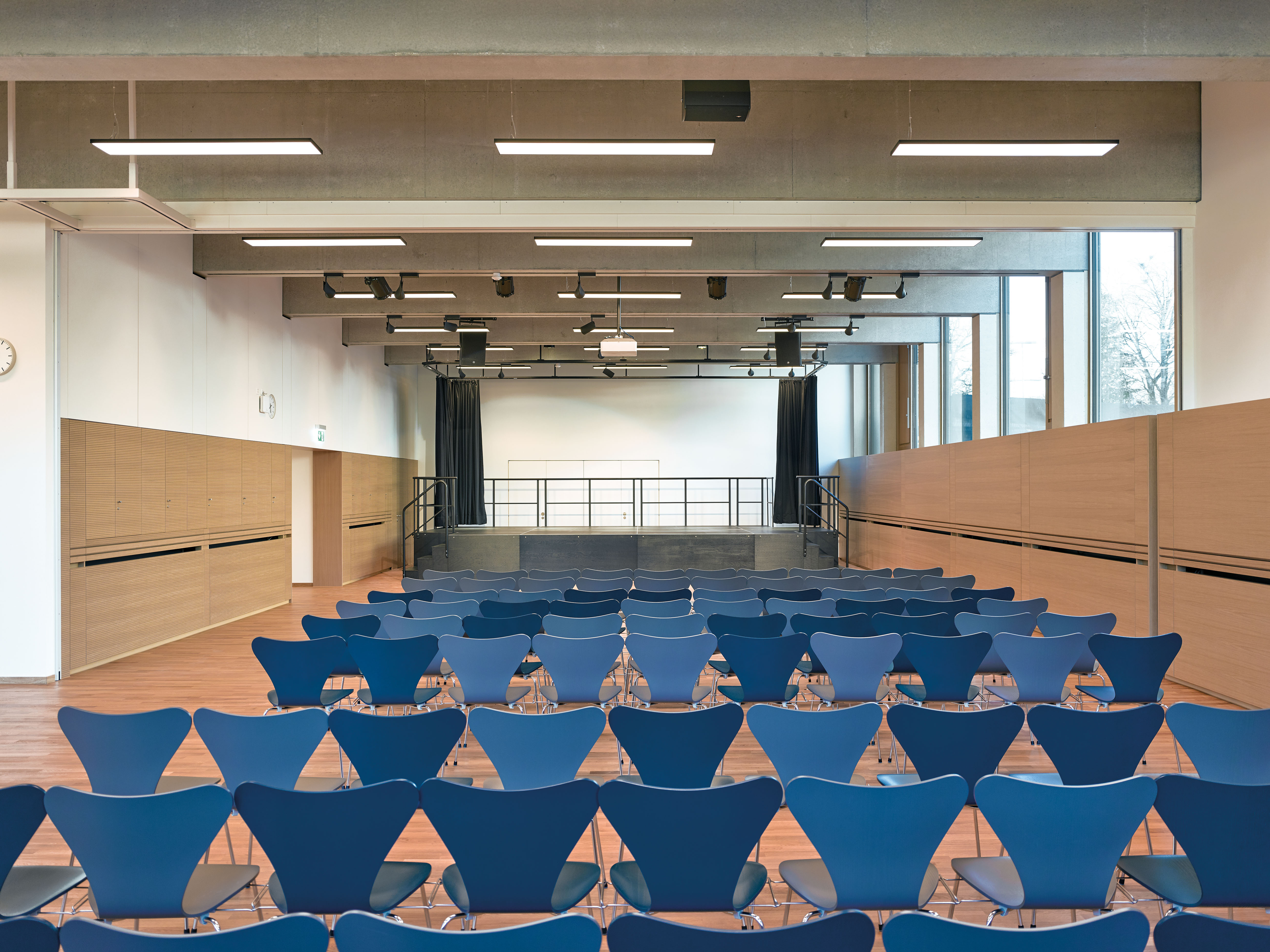 School assembly hall with rows of blue chairs, wood paneling and a curtained stage