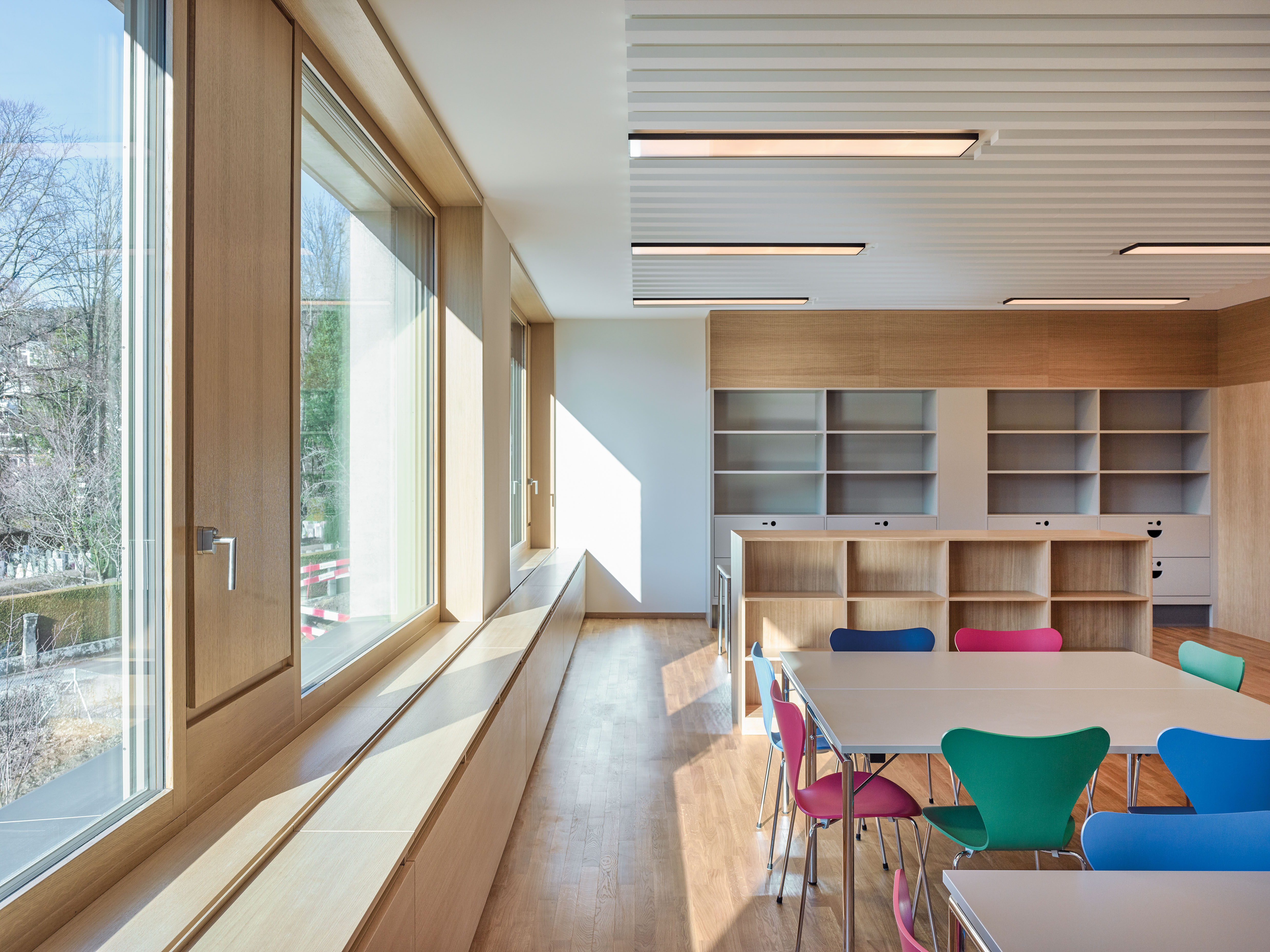 Sunlit school classroom with wood paneling, built-in shelving and colourful chairs around a table