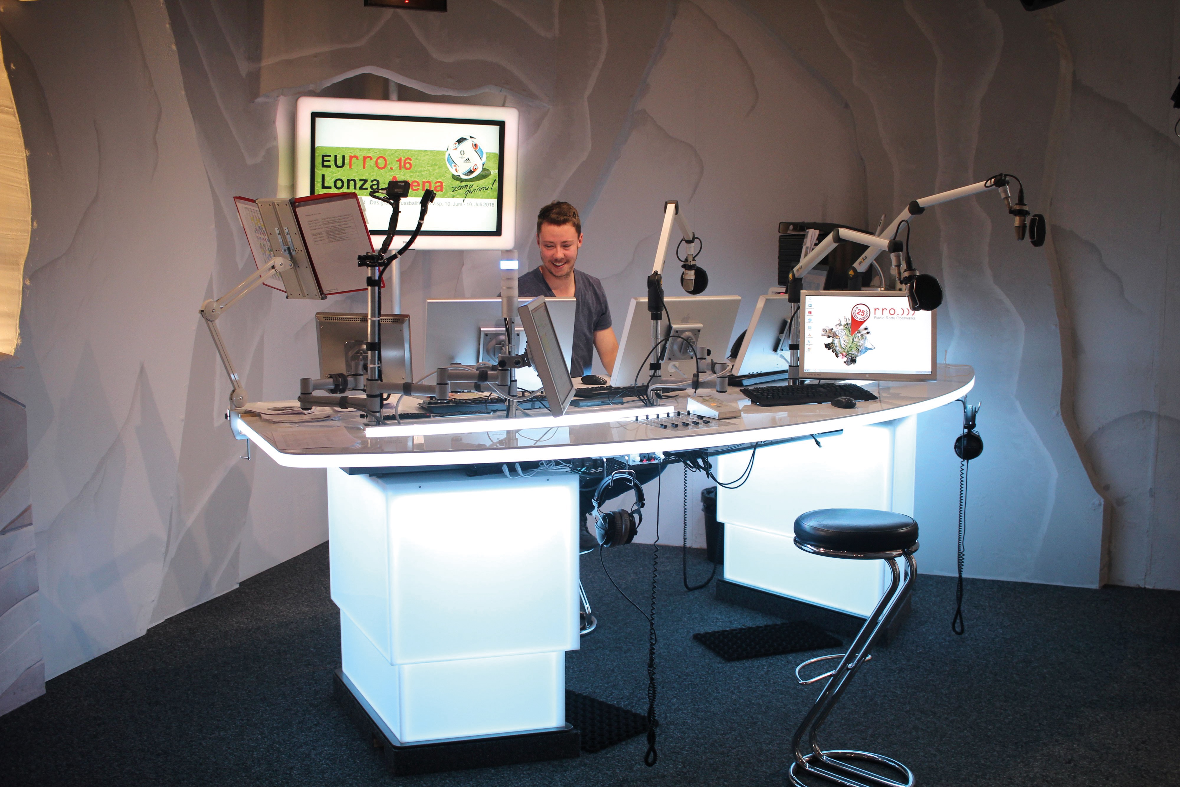 Presenter at the illuminated broadcast desk of Radio Rottu with mic arms and monitors