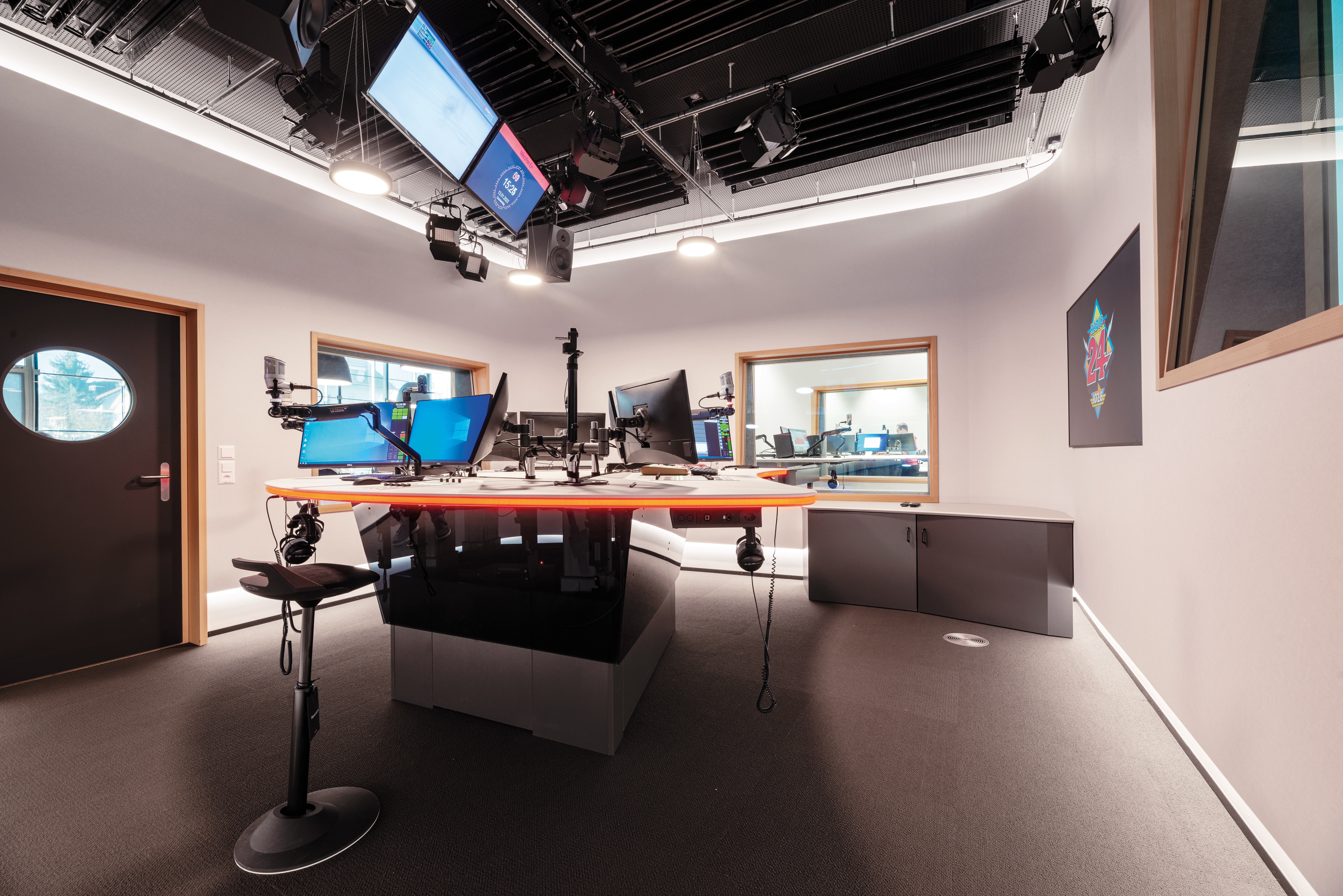Broadcast studio with an orange curved control desk, mic arms and multiple screens