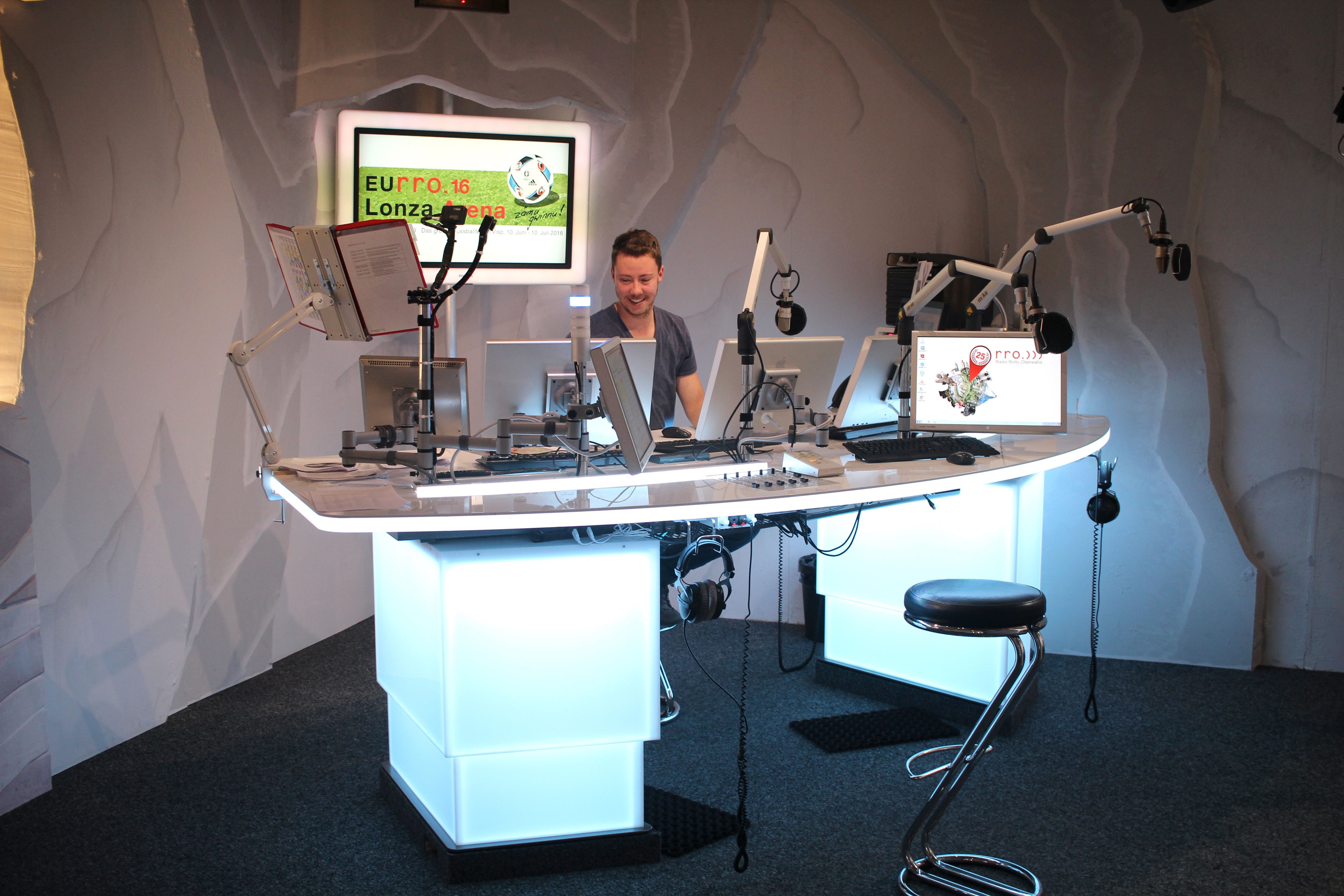 Curved, illuminated white broadcast desk with monitors and microphone arms in the Radio Rottu studio