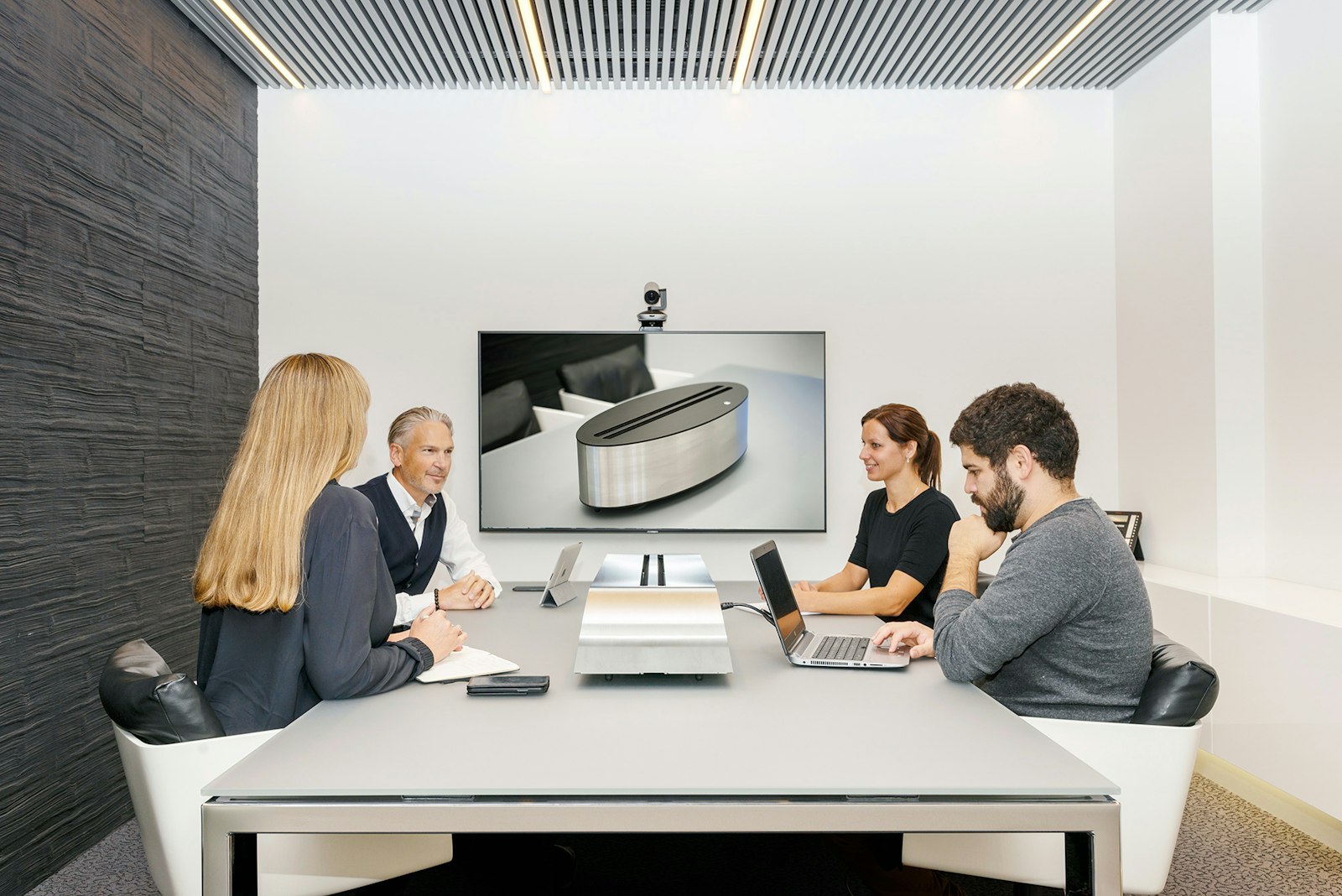 Four people in a meeting room with a video-conferencing screen and laptops