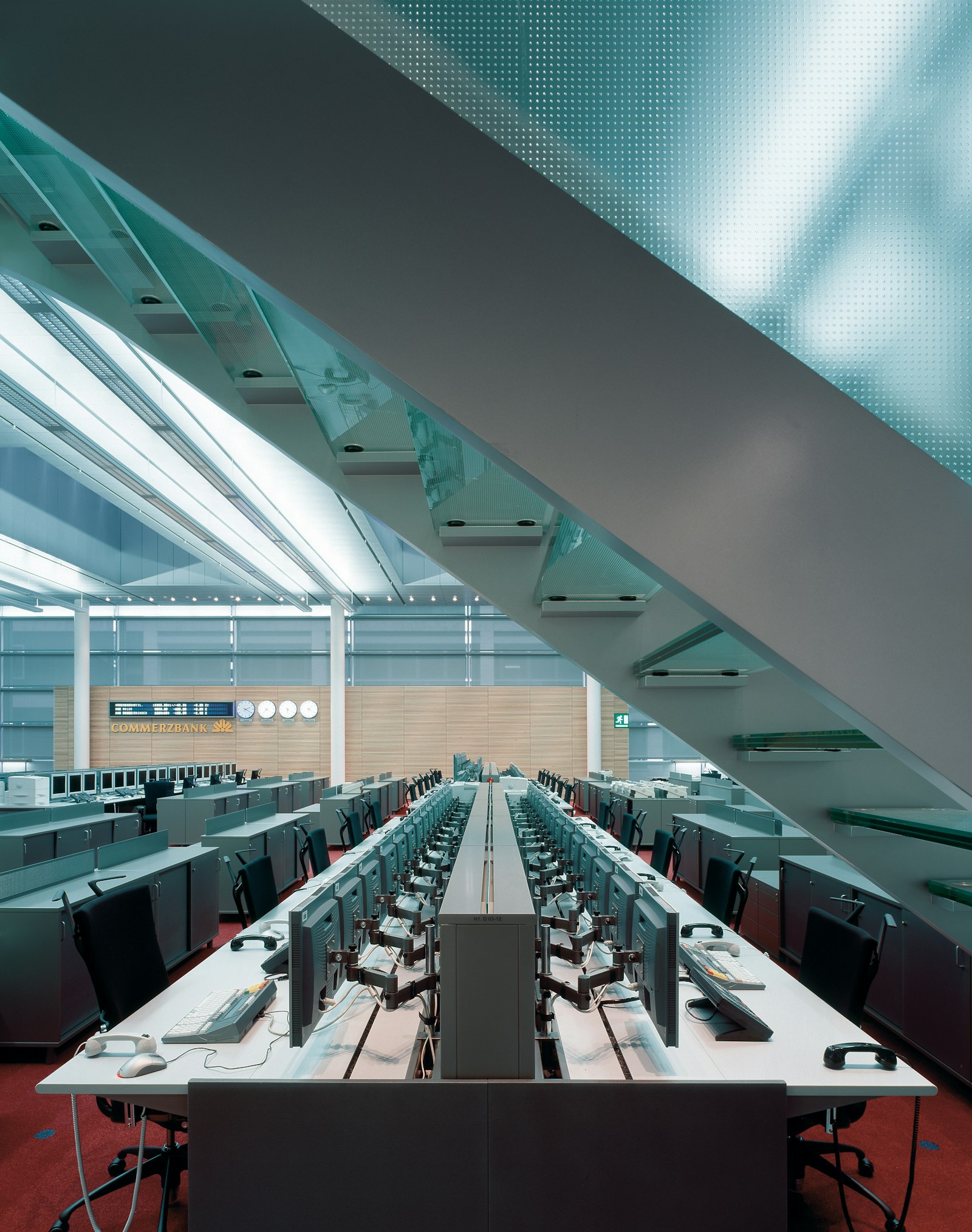 Trading floor with long rows of desks beneath a glass staircase, Commerzbank signage in the background