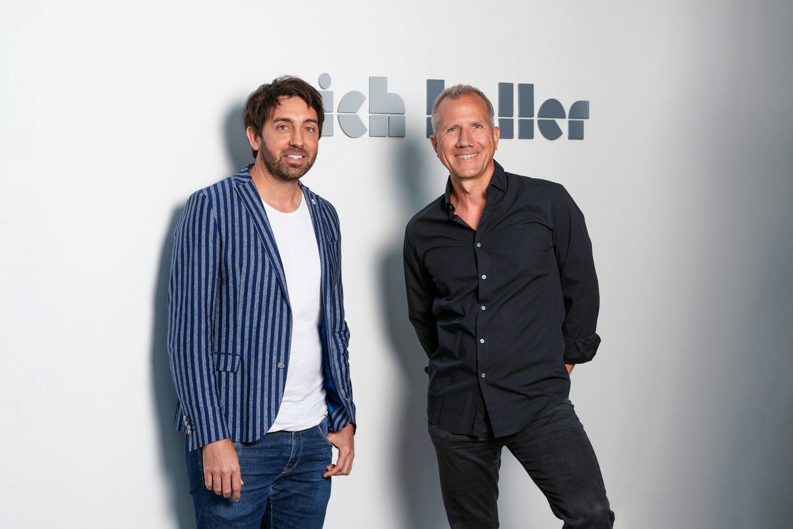 Two men stand smiling in front of the Erich Keller logo on the wall, milestone 2025