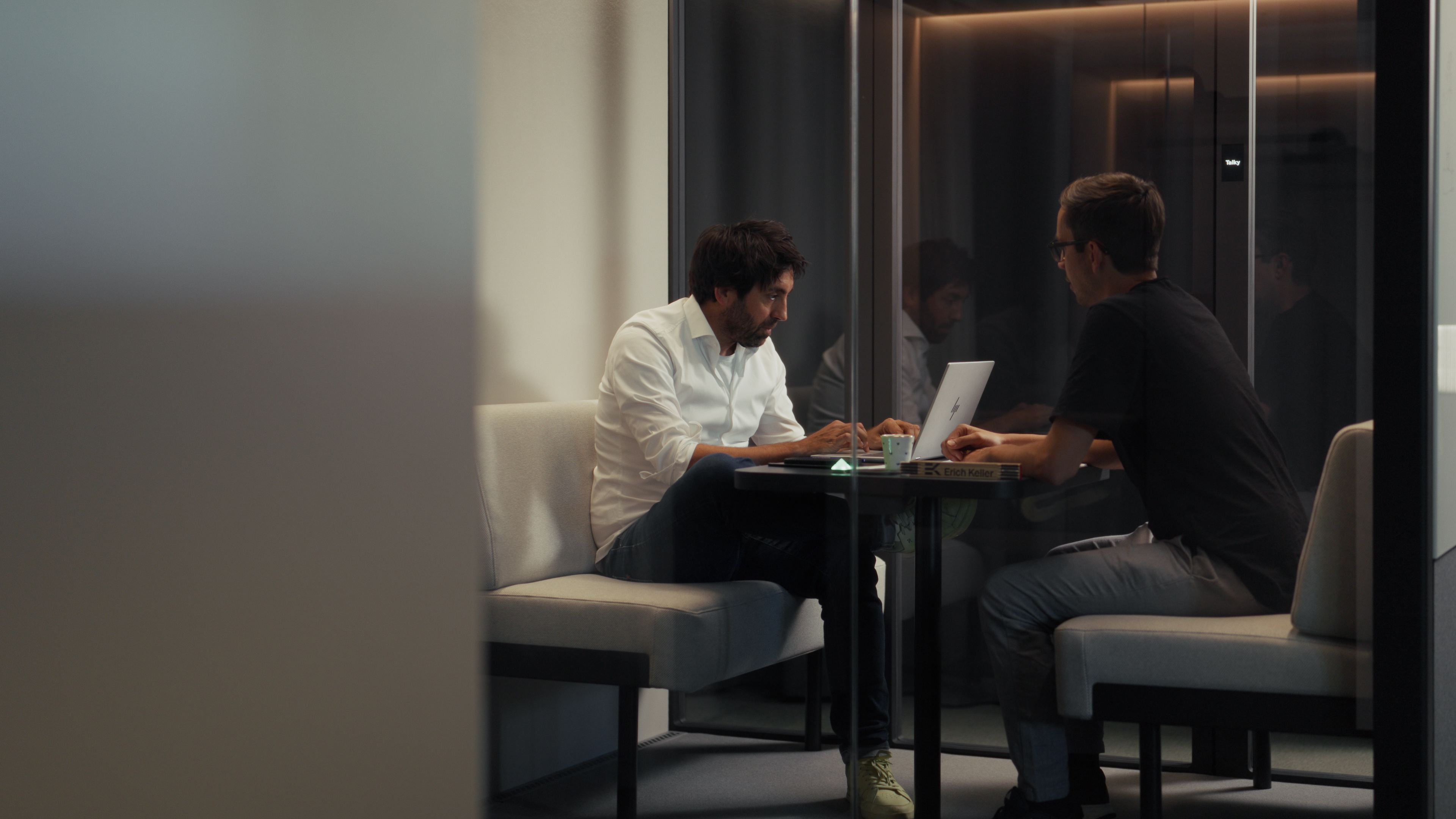 Zwei Kollegen besprechen sich bei Kaffee an einem Laptop in einer Talky-Lounge-Kabine
