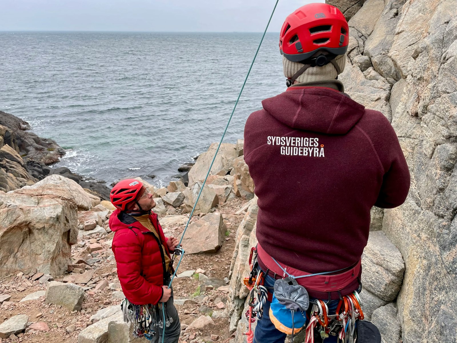 Klätterinstruktör med texten Sydsveriges Guidebyrå på ryggen övervakar topprepssäkring.