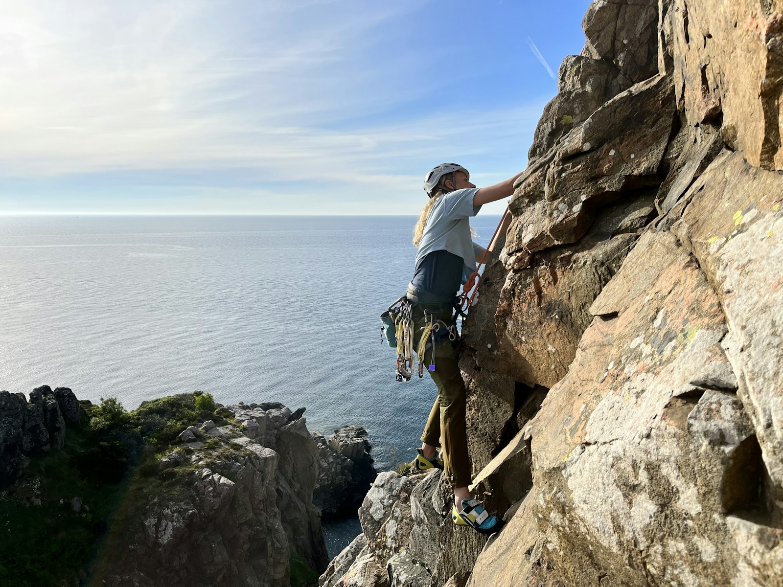 En person klädd i klättringsutrustning klättrar uppför en klippa vid havet, under en delvis molnig himmel, med vatten och fler klippor synliga i bakgrunden.