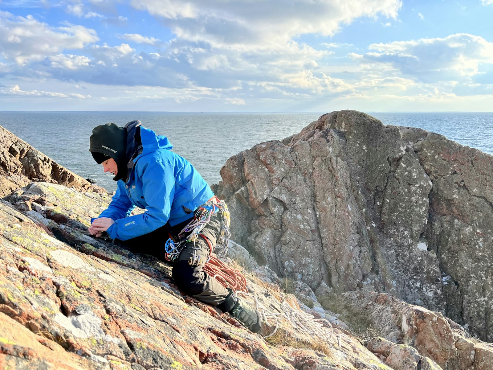 En person i blå jacka och klätterutrustning klättrar uppför en stenig, sluttande klippa vid havet under en delvis molnig himmel.