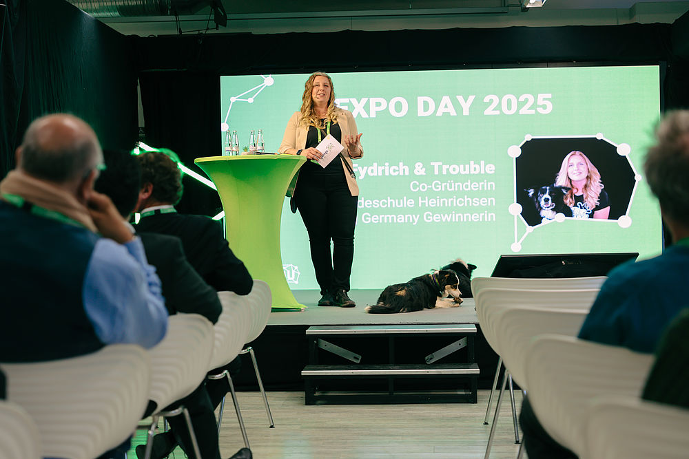 Eine Frau spricht auf der Bühne beim EXPO DAY 2025, neben ihr sitzt ein Hund, und das Publikum schaut ihr zu. Auf einem großen Bildschirm im Hintergrund sind ihr Foto mit dem Hund sowie Veranstaltungsdetails auf Deutsch zu sehen.