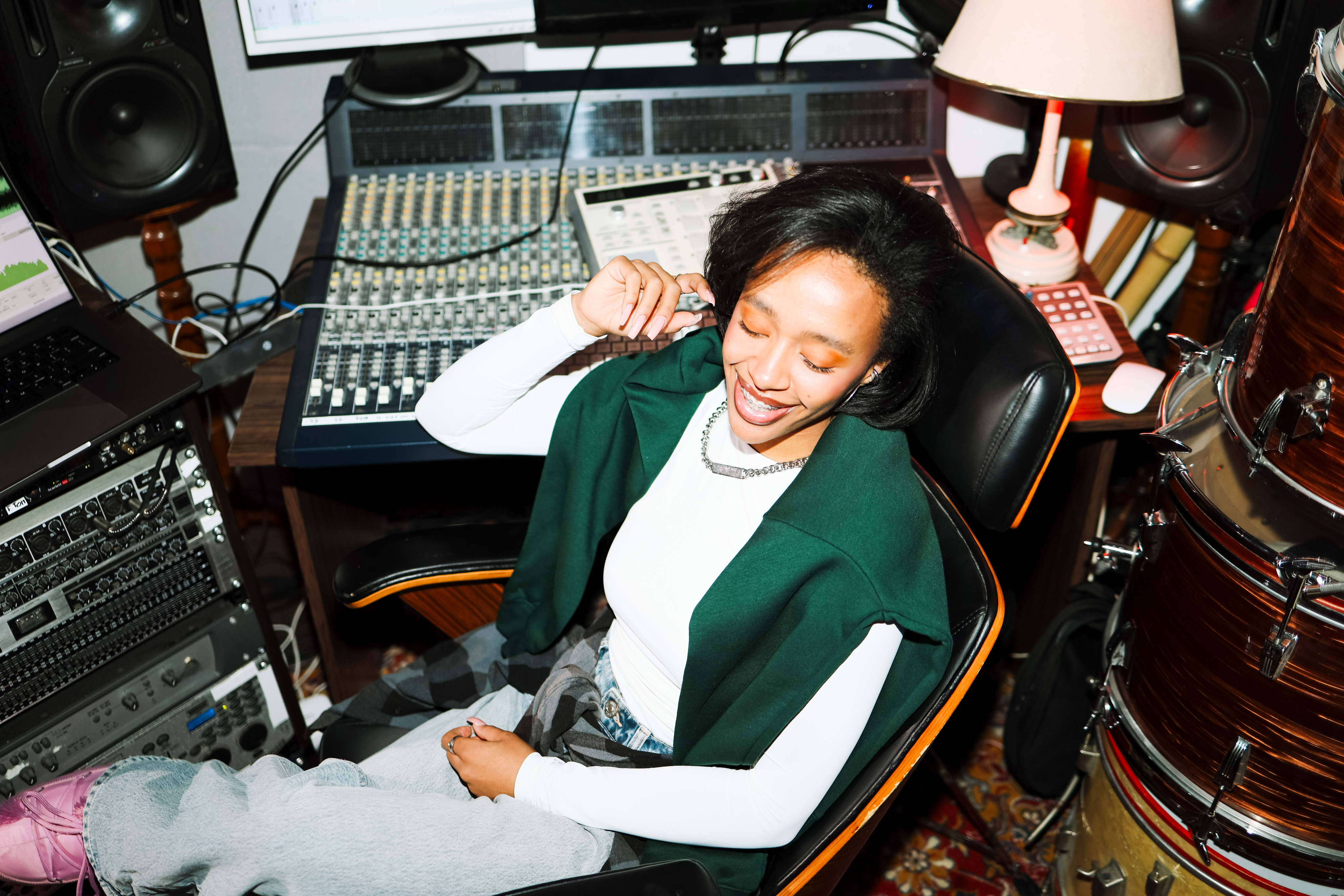 A woman sitting in a music studio environment. Behind her is a mixing board, a studio rack with mountable audio hardware, studio monitors, and a laptop. To the right of her, a stack of drums for a drum kit is visible