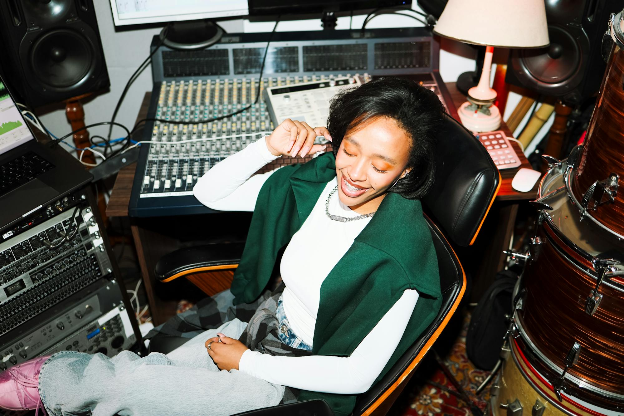 A woman sitting in a music studio environment. Behind her is a mixing board, a studio rack with mountable audio hardware, studio monitors, and a laptop. To the right of her, a stack of drums for a drum kit is visible