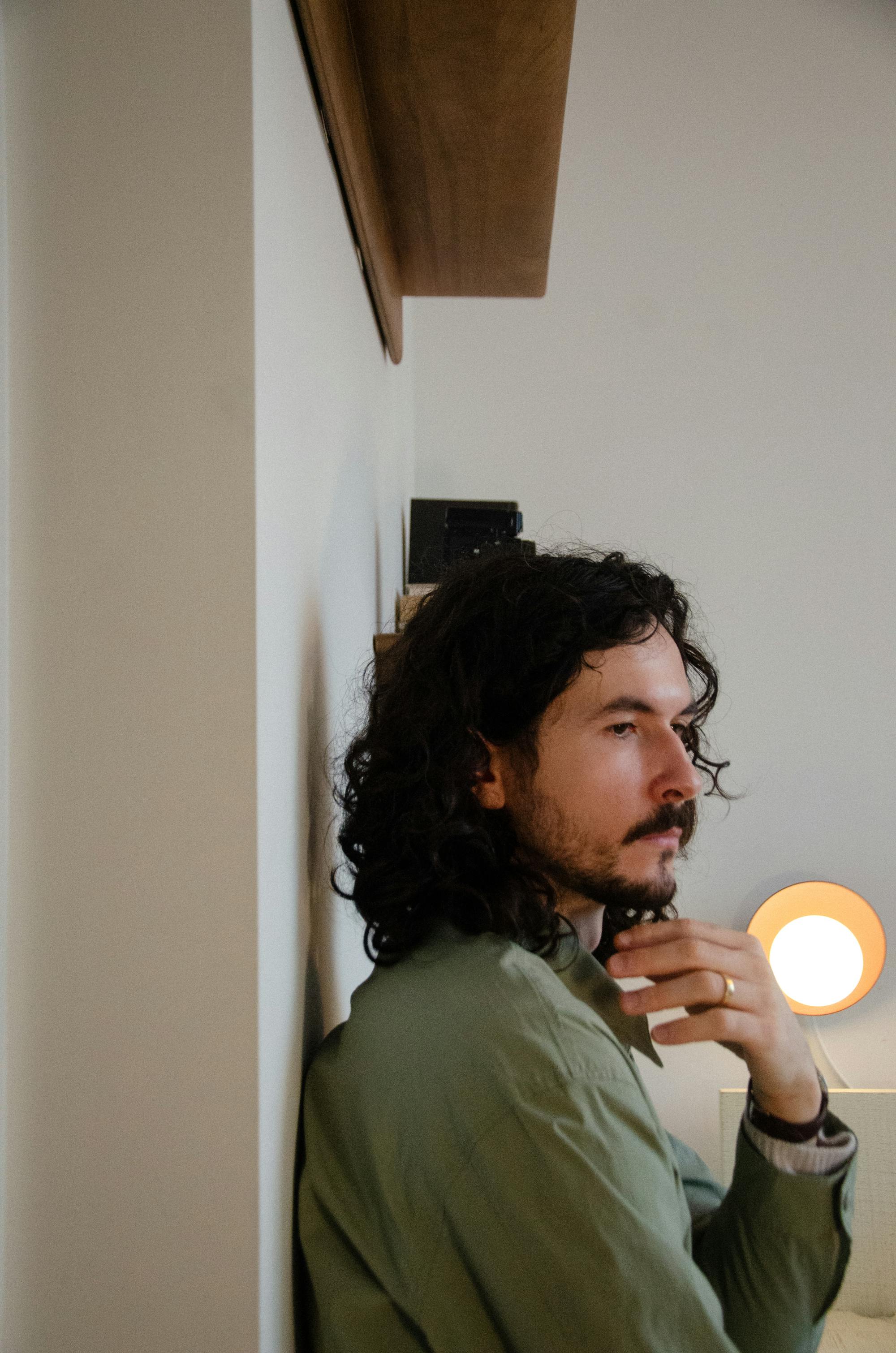 Side profile view of Chris Mazuera leaning against a white wall. A wooden shelf is visible above him and a small lamp is visible in the background.