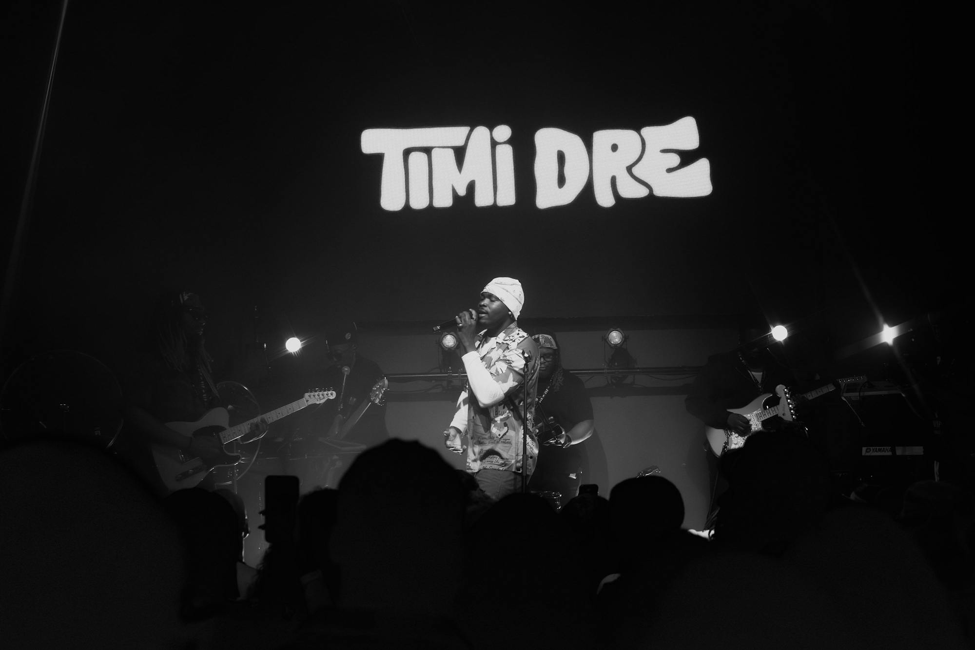 Timi Dre performing live with a band on stage in front of a crowd covered in shadows
