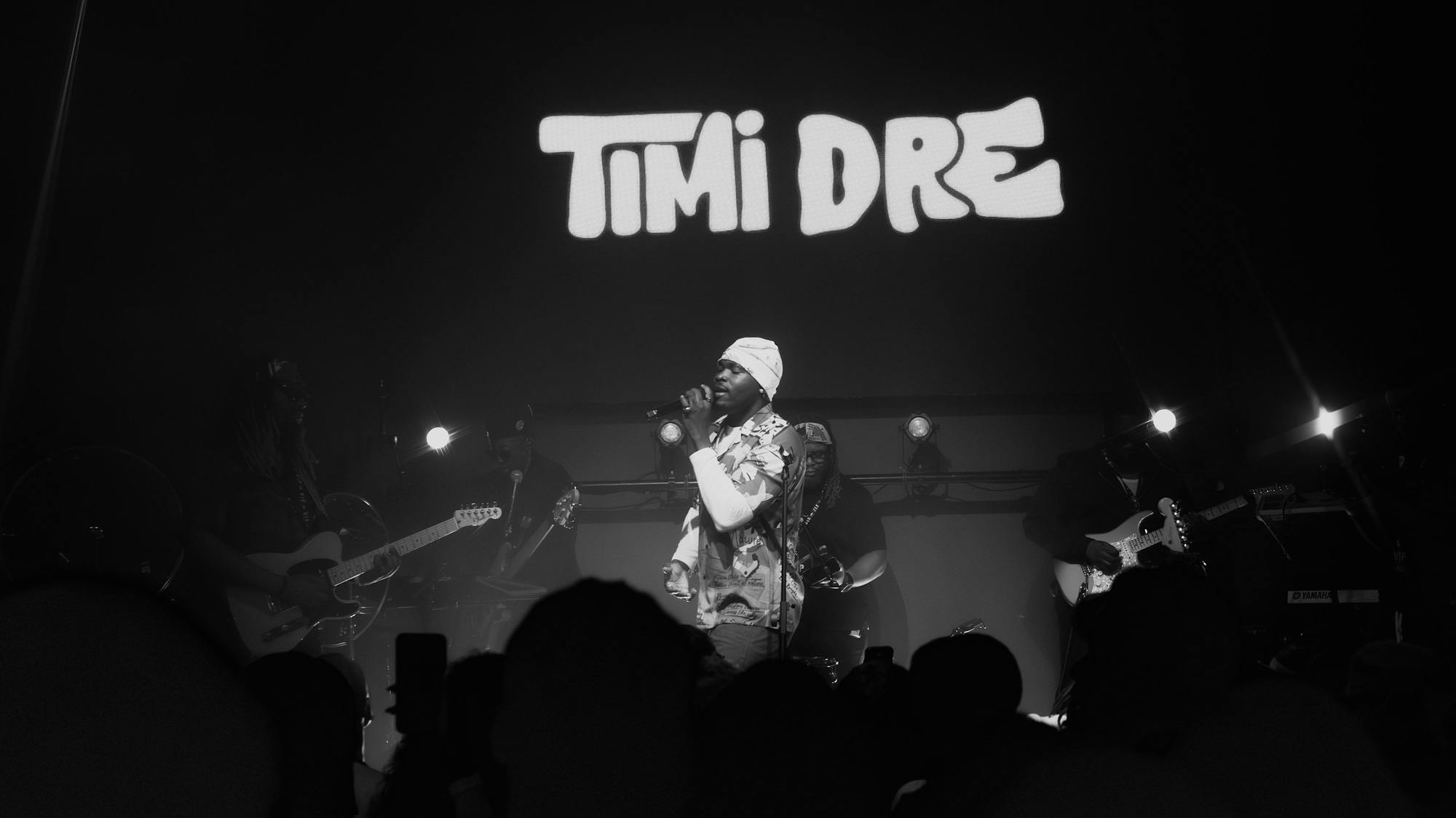 Timi Dre performing live with a band on stage in front of a crowd covered in shadows