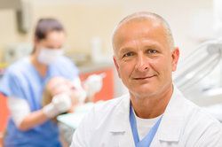 A dentist smiling, with a patient undergoing a dental cleaning in the background