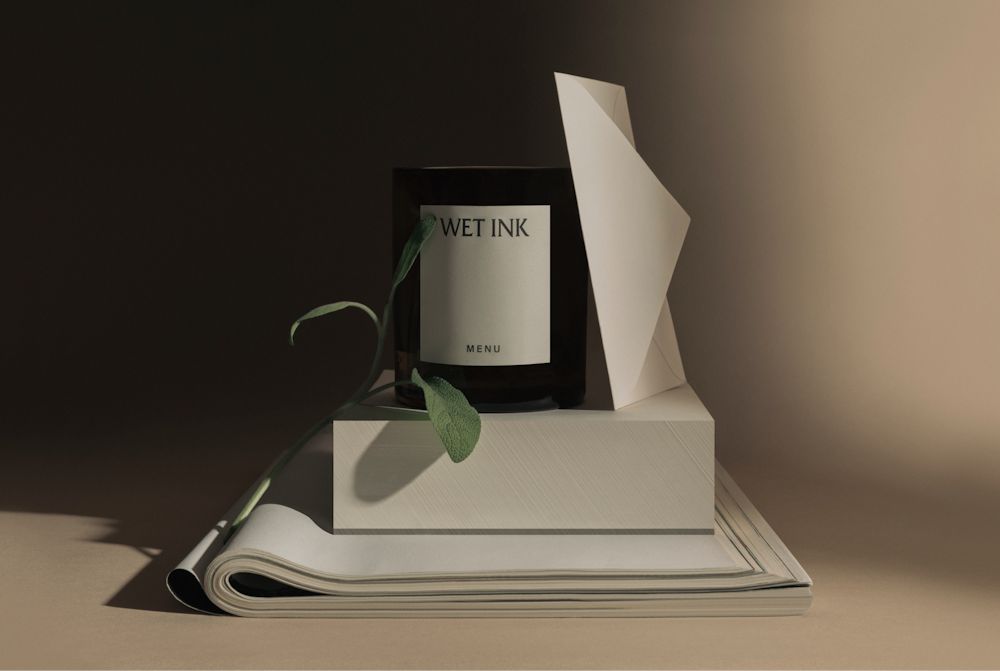 A Menu candle "Wet Ink" on a pile of paper, enveloppe and plant. Elegant light brown background.