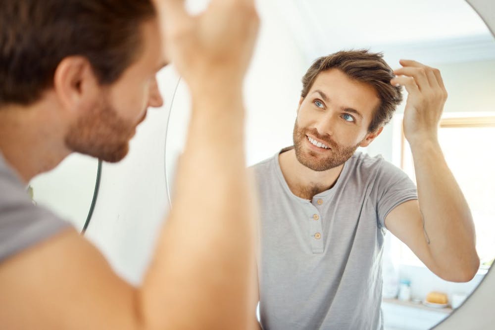 Optimistic man inspecting new hair growth in mirror, hinting at stem cell hair restoration benefits.