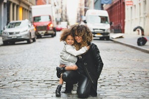 Mini me acquired A woman in a black outfit kneels on a cobblestone street, hugging a smiling child wearing a silver jacket. Vehicles line the urban scene, conveying warmth and happiness.