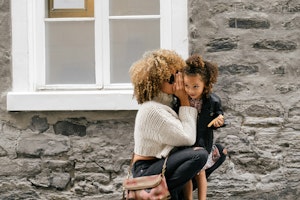 Top secret intel transfer A woman in a cozy sweater crouches to whisper to a smiling young girl in a jacket against a stone wall. The scene conveys warmth and secrecy.