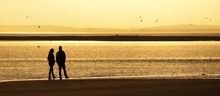 A couple walk along the sand at  Bill Point beach during low tide as the sun sets.  The Isle of Wight can be seen in the background and sea birds flying overhead 