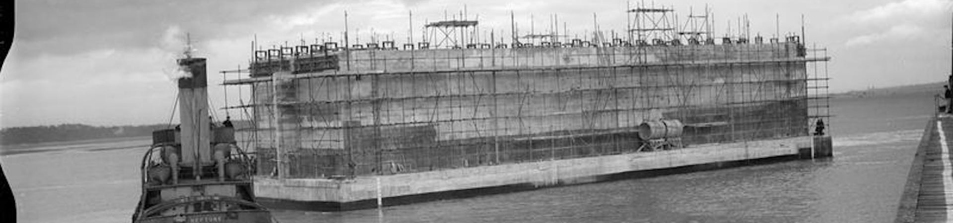 Black and white image of the huge concrete structure of the Phoenix Caison being towed by a tug