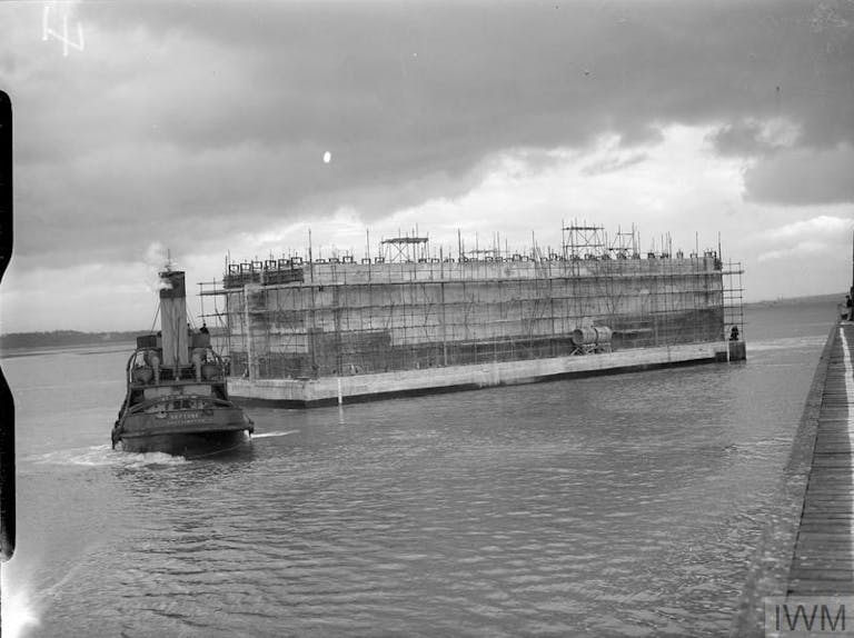 Flouting Phoenix Caisons Pulled by a Tug, courtesy of IWM Black and white image of the huge concrete structure of the Phoenix Caison being towed by a tug