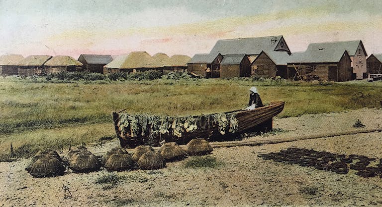 Old image of a fisherman drying out his nets over his rowing boat with willow lobster pots drying on the sand in the foreground.  In the background are the fishermen's huts at East Beach, Selsey. 