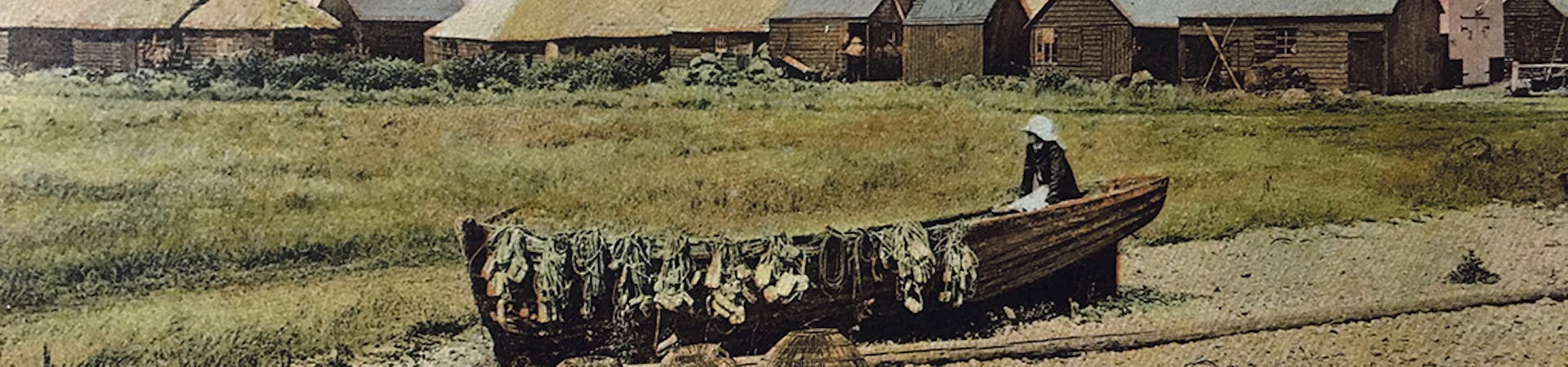 Old image of a fisherman drying out his nets over his rowing boat with willow lobster pots drying on the sand in the foreground. In the background are the fishermen's huts at East Beach, Selsey.