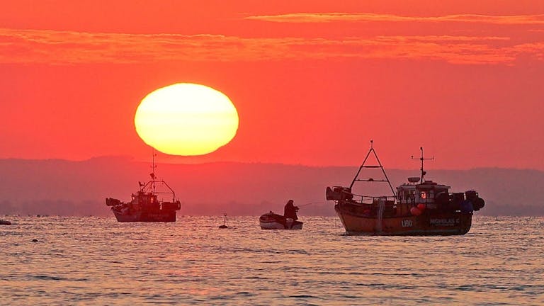 Lone fisherman sandwiched on his dingey with fishing rod, between two larger fishing boats at sunrise 