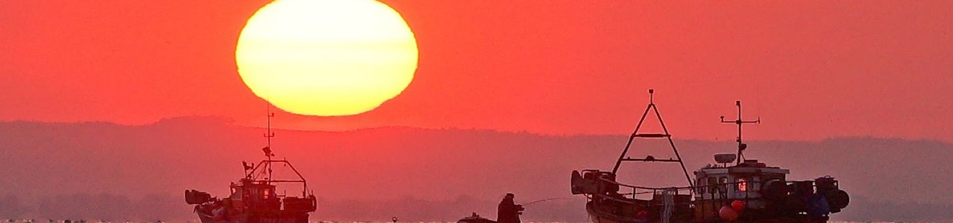 Lone fisherman sandwiched on his dingey with fishing rod, between two larger fishing boats at sunrise