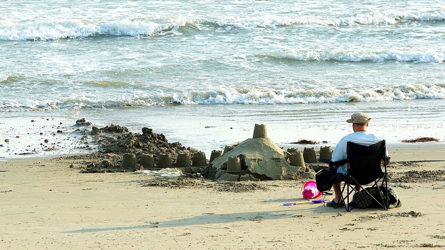 Sandcastles being taken by the incoming tide on Selsey Oval Beach with a man sat in a chair relaxing, facing the sea with a pink sandcastle bucket at his feet. 