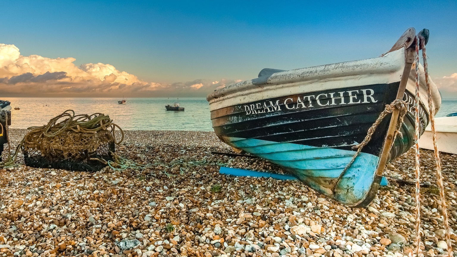 A dark blue with light blue bottom boat called Dream Catcher on the shingle at east beach with a lobster pot to its left.  The clouds are creating cotton wool white shapes within the blue sky as the sun captures the sea water 