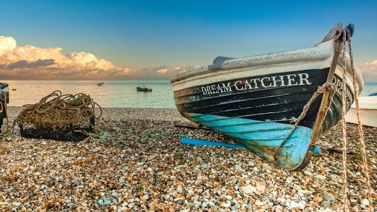 A dark blue with light blue bottom boat called Dream Catcher on the shingle at east beach with a lobster pot to its left.  The clouds are creating cotton wool white shapes within the blue sky as the sun captures the sea water 
