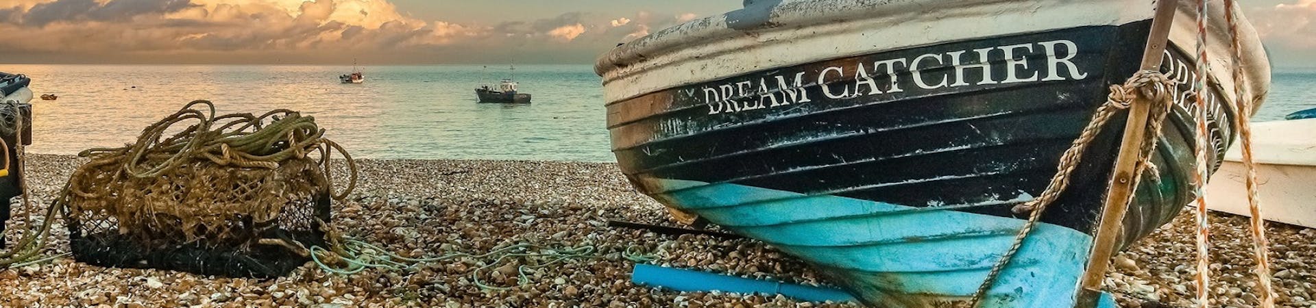 A dark blue with light blue bottom boat called Dream Catcher on the shingle at east beach with a lobster pot to its left. The clouds are creating cotton wool white shapes within the blue sky as the sun captures the sea water