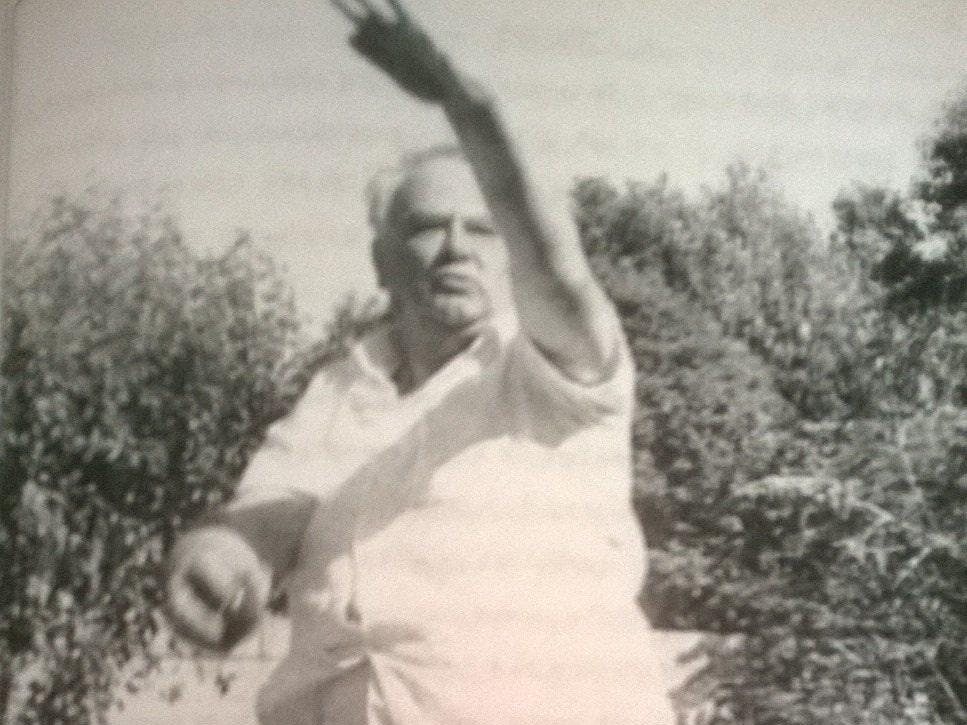 A black and white image of Sir Patrick Moore wearing cricket whites and in mid bowl action