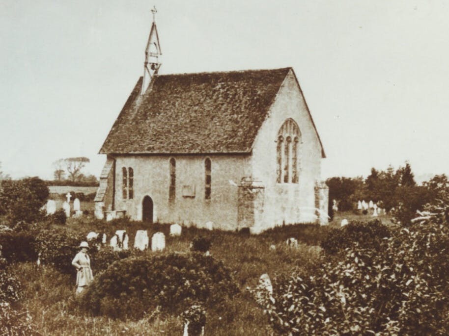 An image of St Wilfrids taken in the early 20th Century in black and white depicting a woman standing in the overgrown graveyard 