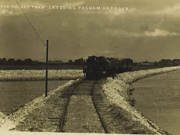 Black and white image showing The Selsey Tram crossing a raised area of land
