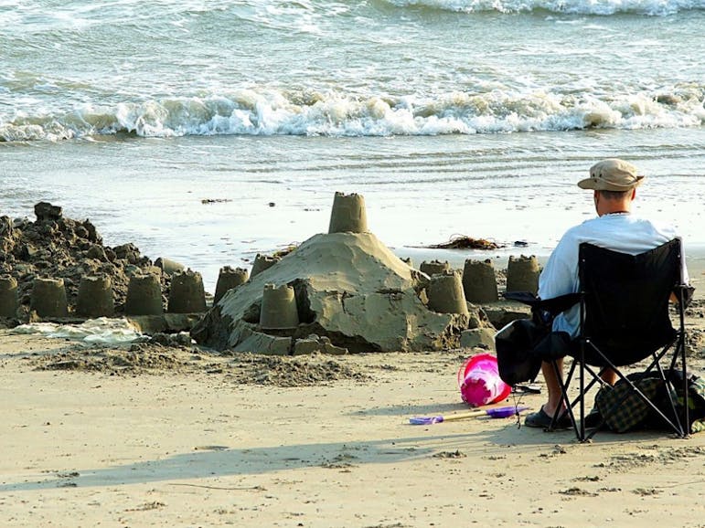 Sandcastle in Selsey Photograph of the back of a man sat in a beach chair, next to a sandcastle with a discarded pink bucket